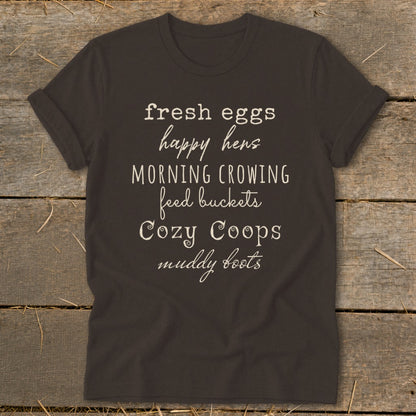 Dark t-shirt with white text listing farm-related phrases: fresh eggs, happy hens, morning crowing, feed buckets, cozy coops, muddy boots.