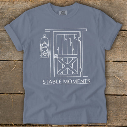 Blue t-shirt with white line drawing of barn door and lantern, text "STABLE MOMENTS".