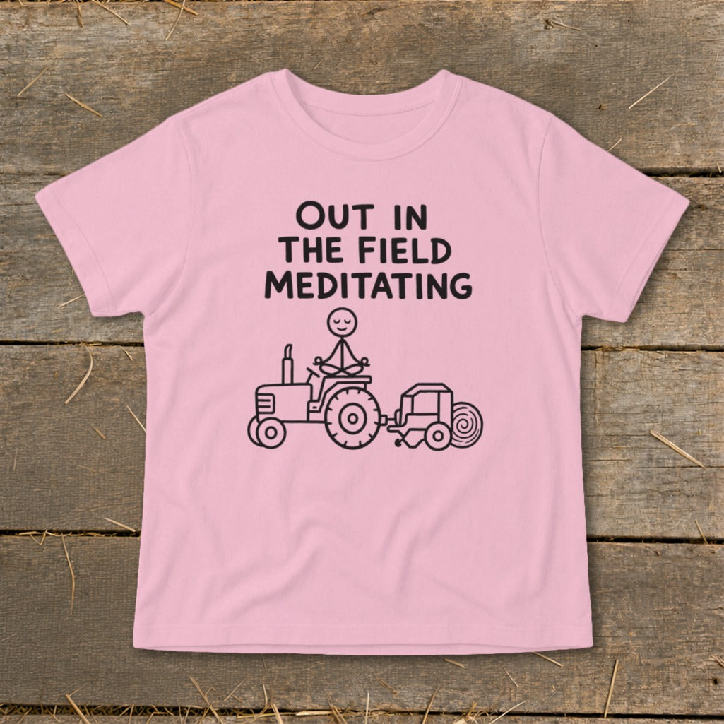 Pink t-shirt with black text "OUT IN THE FIELD MEDITATING" and drawing of stick figure meditating on tractor.