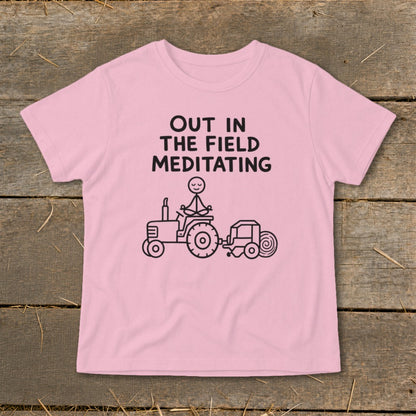 Pink t-shirt with black text "OUT IN THE FIELD MEDITATING" and drawing of stick figure meditating on tractor.