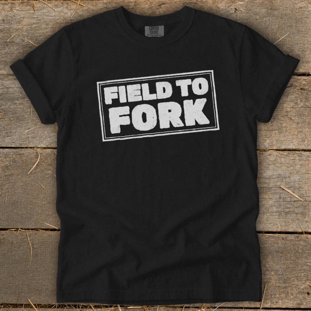 A black t-shirt with rolled-up sleeves, featuring the distressed white text FIELD TO FORK centered on the chest, lying flat on a wooden background.