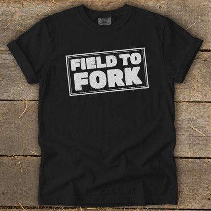 A black t-shirt with rolled-up sleeves, featuring the distressed white text FIELD TO FORK centered on the chest, lying flat on a wooden background.