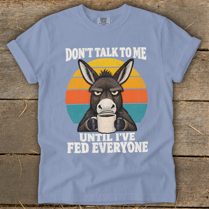 A blue t-shirt featuring a graphic of a grumpy donkey drinking coffee with sunset stripes and text.