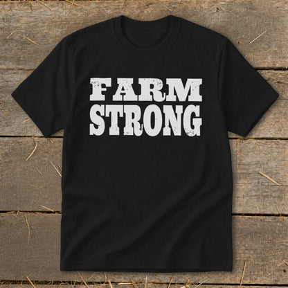 Black t-shirt featuring distressed white block letters that read FARM STRONG, laid flat on a rustic wooden background.