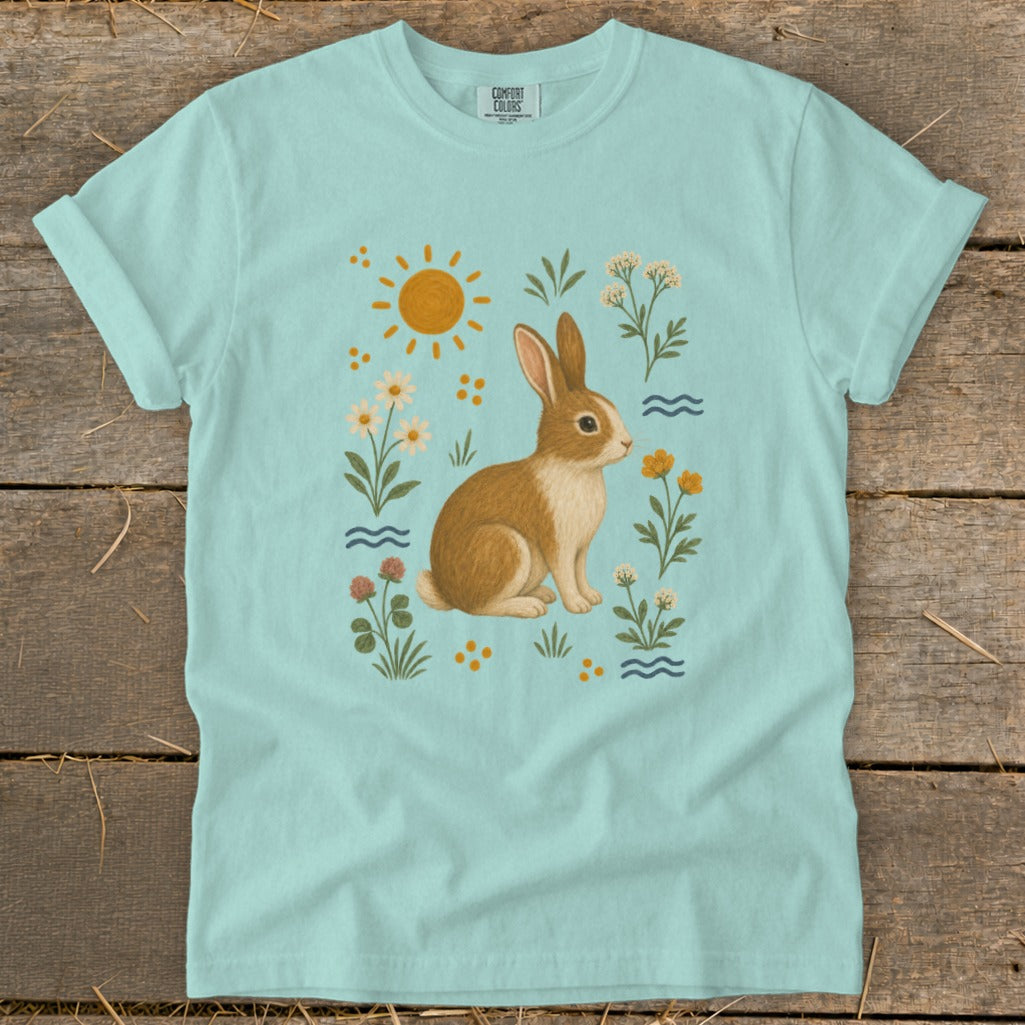 Light blue t-shirt with a watercolor illustration of a brown and white rabbit surrounded by flowers, sun, and waves.
