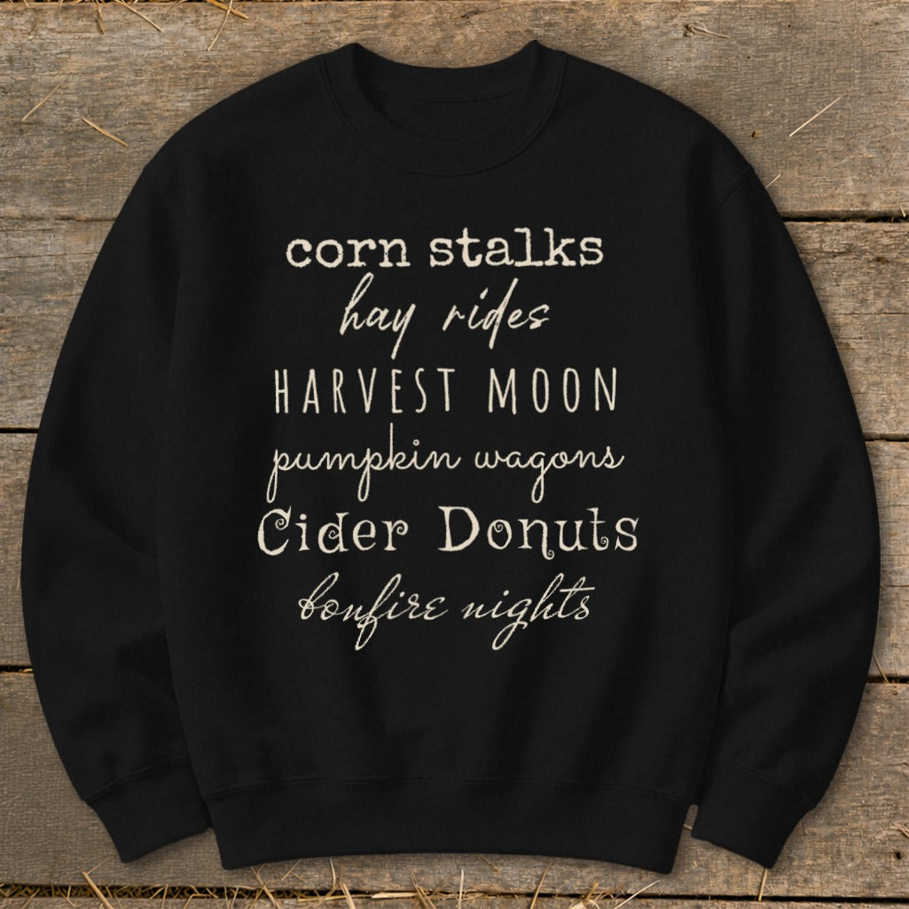 Black sweatshirt with fall-themed text: corn stalks, hay rides, harvest moon, pumpkin wagons, cider donuts, bonfire nights.