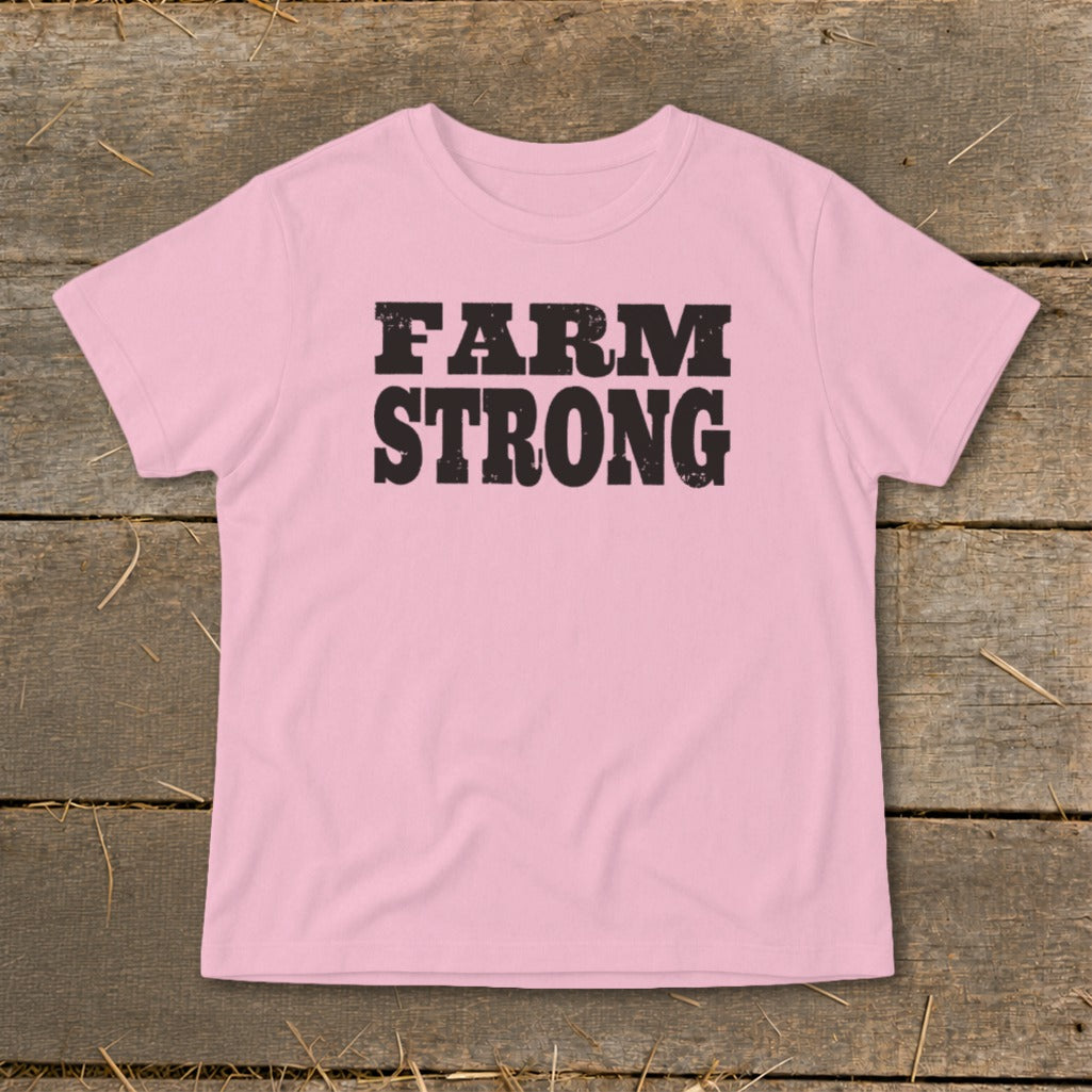 A light pink short-sleeved t-shirt with the text FARM STRONG printed in black across the chest, laid flat on a rustic wooden background.