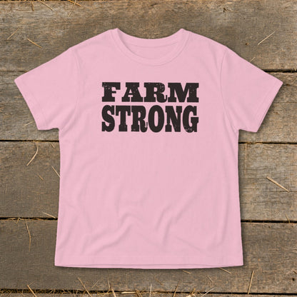 A light pink short-sleeved t-shirt with the text FARM STRONG printed in black across the chest, laid flat on a rustic wooden background.