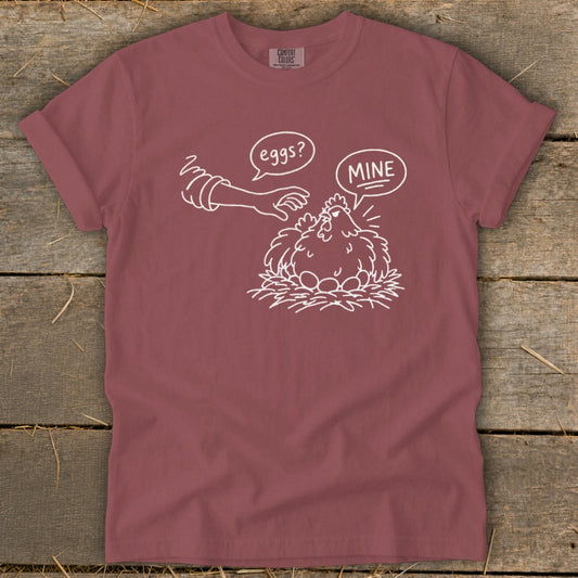 T-shirt with white line drawing of a chicken protecting its eggs, saying "MINE".