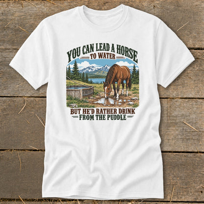 A white t-shirt featuring a graphic of a horse drinking from a puddle with mountains and the text "You can lead a horse to water, but he'd rather drink from the puddle."