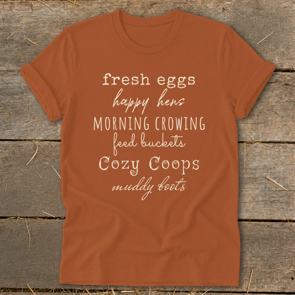 Orange t-shirt with white text: "fresh eggs, happy hens, MORNING CROWING, feed buckets, Cozy Coops, muddy boots."