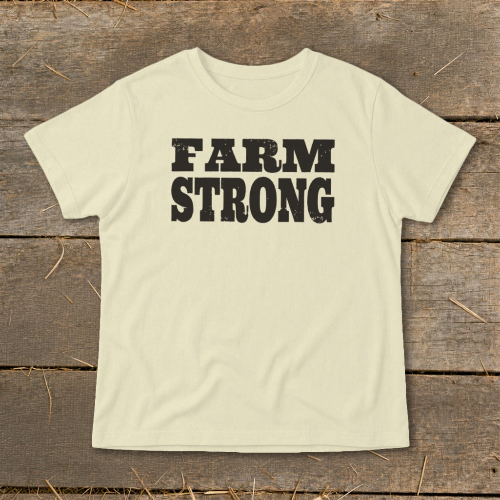 A cream-colored short-sleeved t-shirt with the text FARM STRONG printed in distressed black lettering across the chest, laid flat on a rustic wooden background.