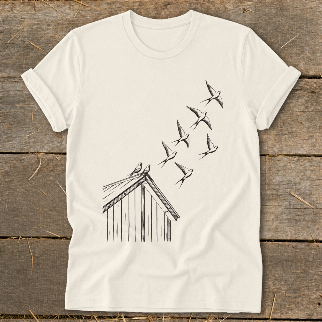 Off-white t-shirt laid flat on wooden planks, featuring a black line art graphic of birds flying from a roof.