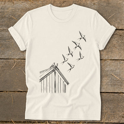 Off-white t-shirt laid flat on wooden planks, featuring a black line art graphic of birds flying from a roof.