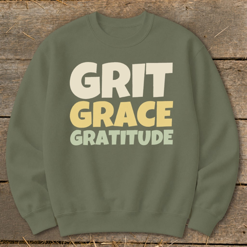 An olive green crewneck sweatshirt featuring the stacked text Grit Grace Gratitude in cream and yellow.