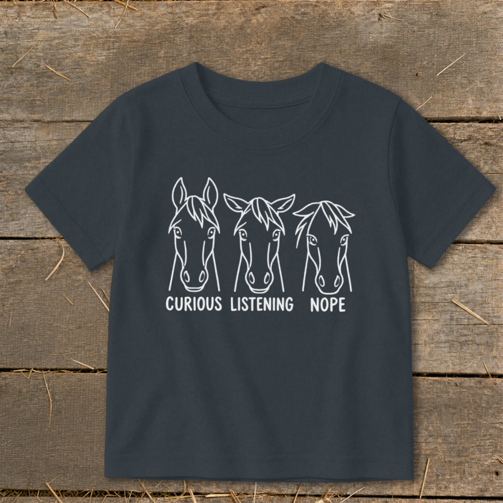 Dark gray children's t-shirt featuring a white graphic of three horse heads with the captions "CURIOUS," "LISTENING," and "NOPE."