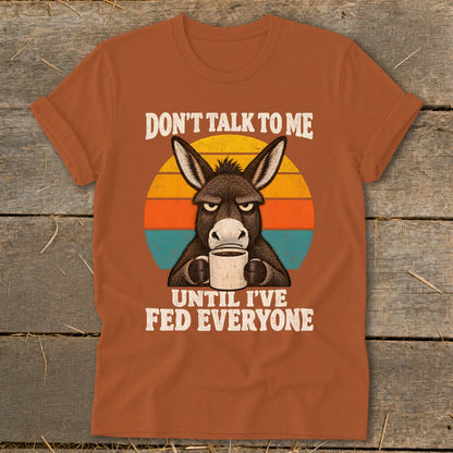 Rust-colored t-shirt featuring a grumpy donkey holding a coffee mug, overlaid on a sunset graphic with text reading, DON'T TALK TO ME UNTIL I'VE FED EVERYONE.