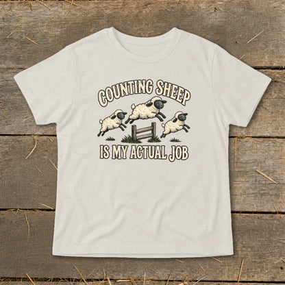 Light beige t-shirt with cartoon sheep jumping a fence, reading "Counting Sheep Is My Actual Job".