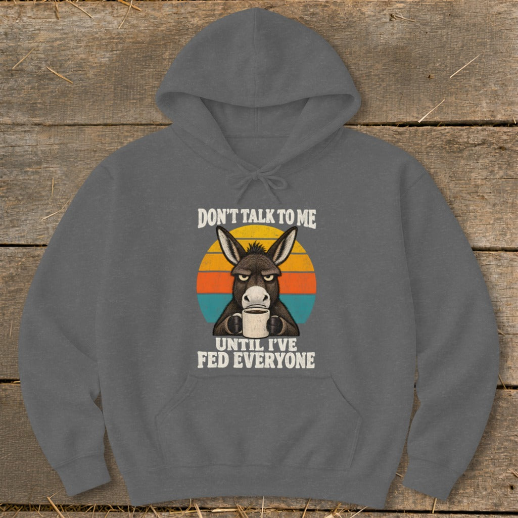 Dark gray hoodie with a graphic of a grumpy donkey holding a coffee mug and text reading "DON'T TALK TO ME UNTIL I'VE FED EVERYONE."