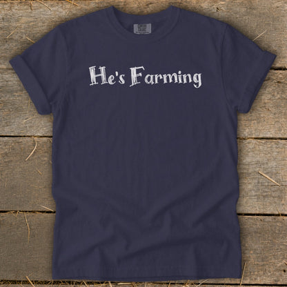 Navy t-shirt with white text "He's Farming" on wooden background.