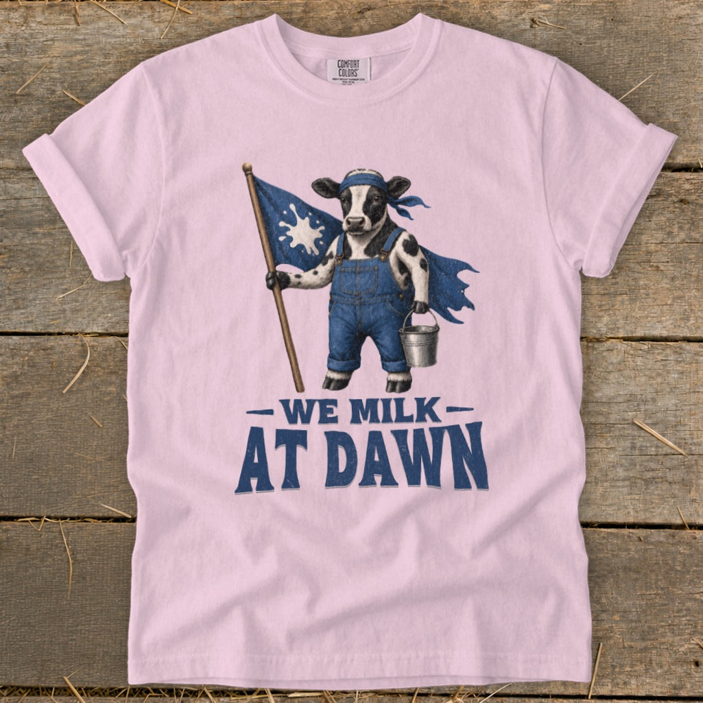 Pink t-shirt graphic of a cow with a cape, flag, and milk pail, reading "WE MILK AT DAWN".