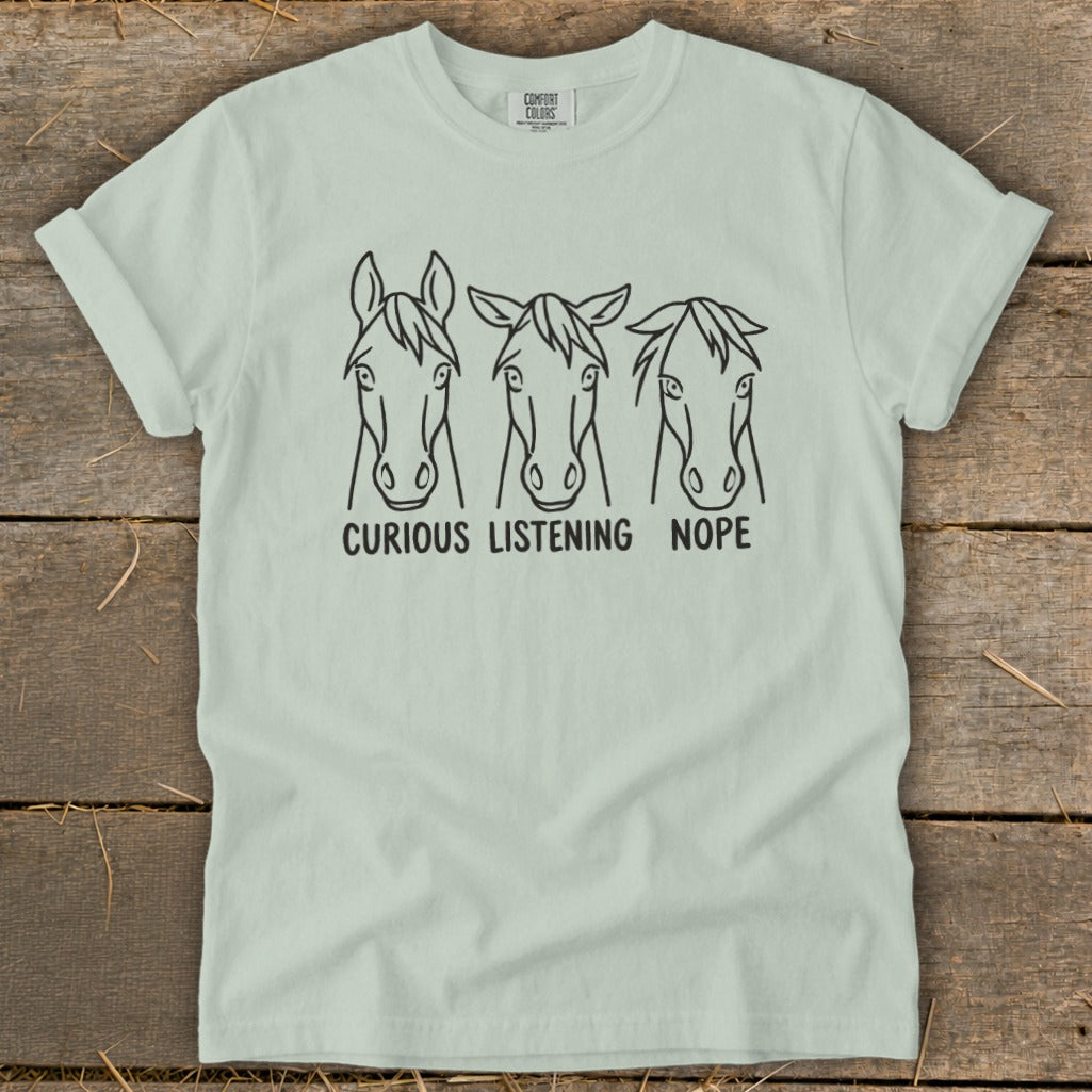 A light sage green t-shirt with three horse head line drawings and text reading Curious, Listening, Nope.