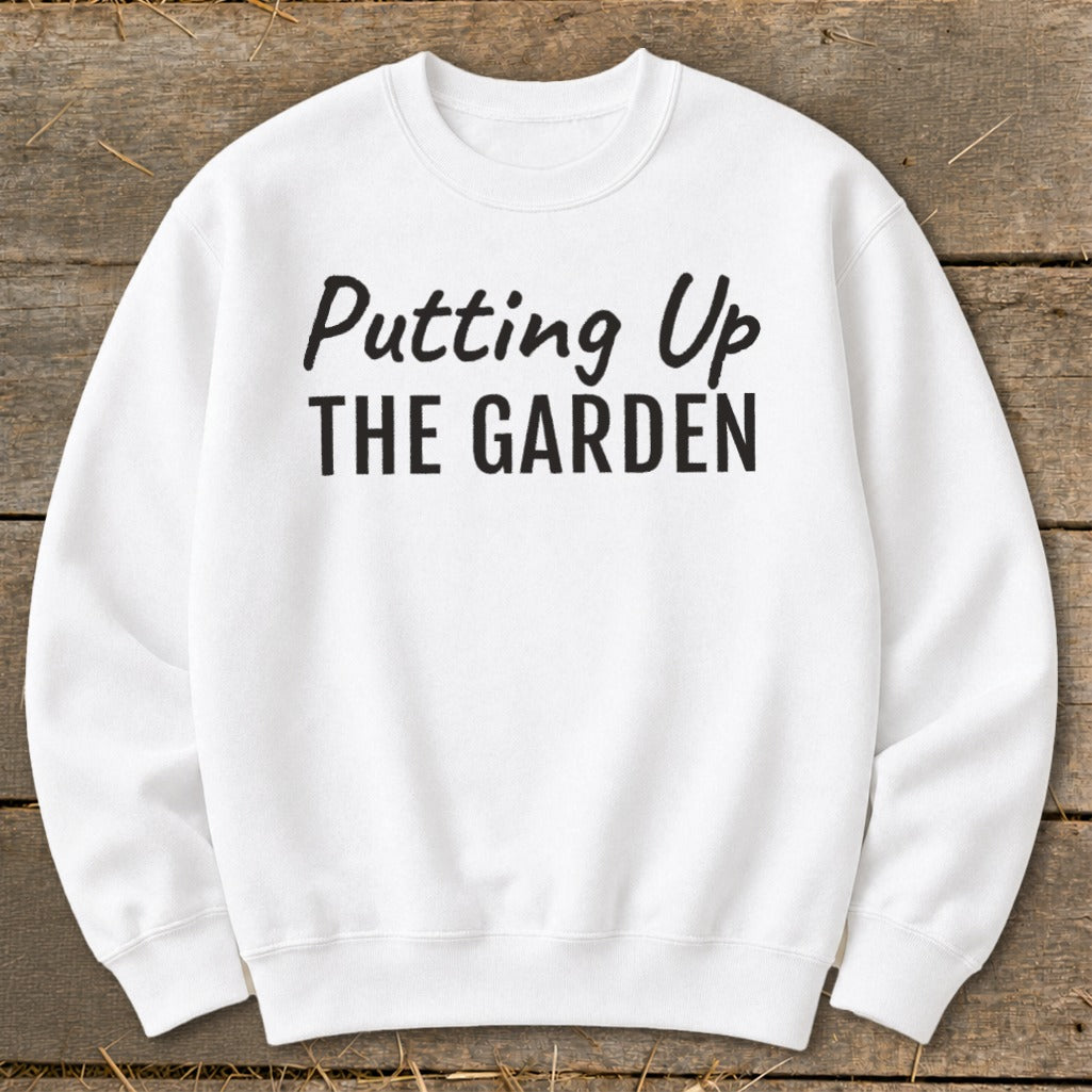 White sweatshirt with black text "Putting Up THE GARDEN" on wooden background.