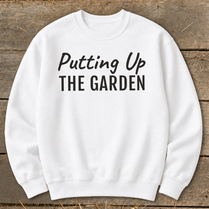 White sweatshirt with black text "Putting Up THE GARDEN" on wooden background.