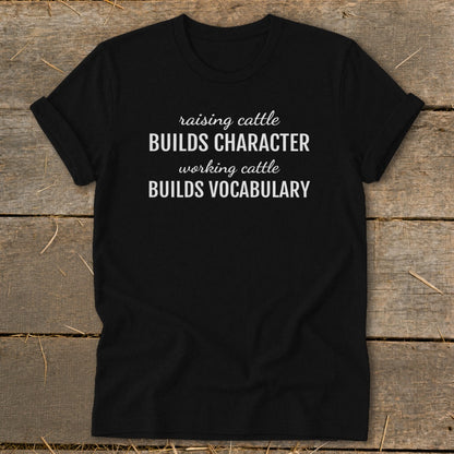 Black t-shirt with white text: "raising cattle BUILDS CHARACTER, working cattle BUILDS VOCABULARY".