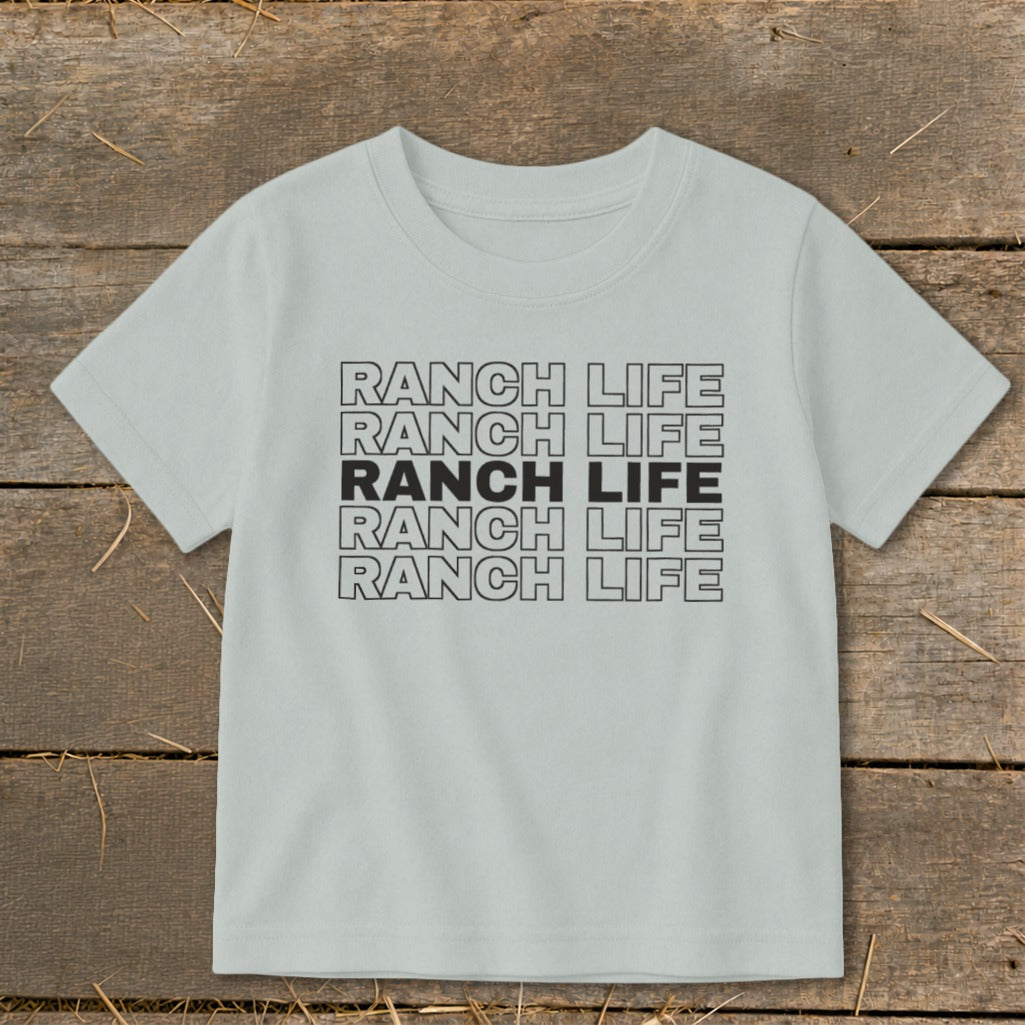 A light gray children's t-shirt printed with stacked repeating text that reads Ranch Life on a wooden background.
