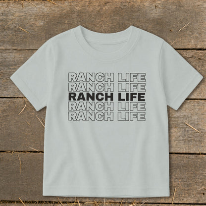 A light gray children's t-shirt printed with stacked repeating text that reads Ranch Life on a wooden background.