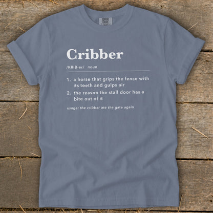 Slate blue crewneck t-shirt featuring dictionary-style text defining the word cribber displayed on a rustic wooden background.