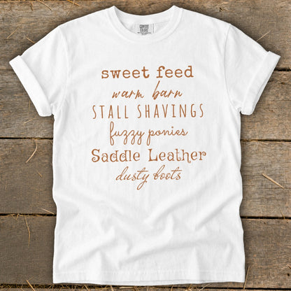 White t-shirt with "sweet feed warm barn stall shavings fuzzy ponies saddle leather dusty boots" text.