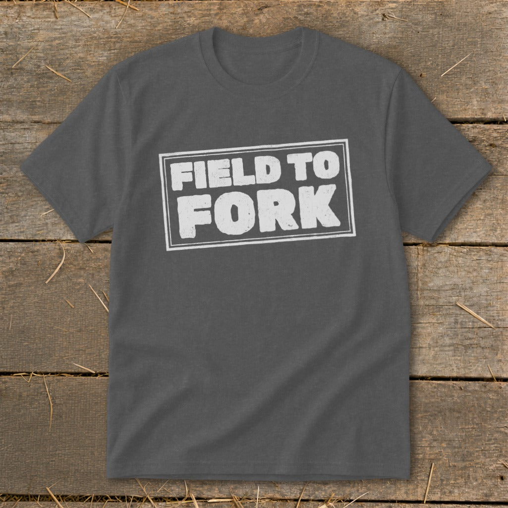 A charcoal gray crewneck t-shirt featuring the white text Field to Fork inside a bordered rectangle, laid flat on a rustic wooden background.