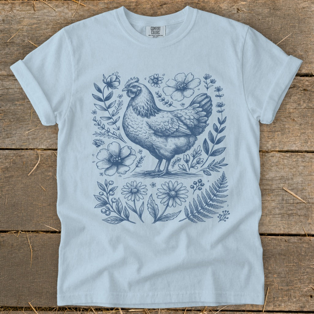 Light blue t-shirt with a blue line drawing of a chicken surrounded by flowers and foliage.