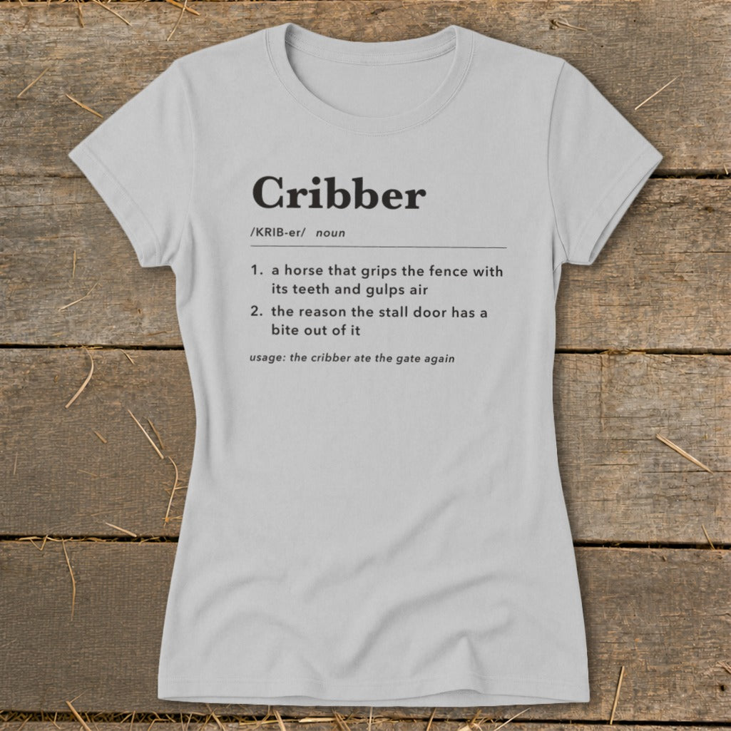 Light gray women's t-shirt printed with a dictionary definition of the word "Cribber" laid flat on wood.