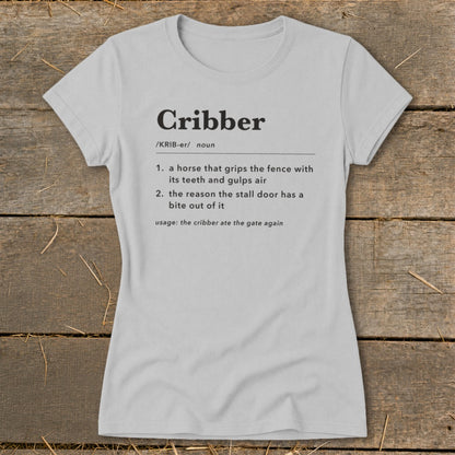 Light gray women's t-shirt printed with a dictionary definition of the word "Cribber" laid flat on wood.