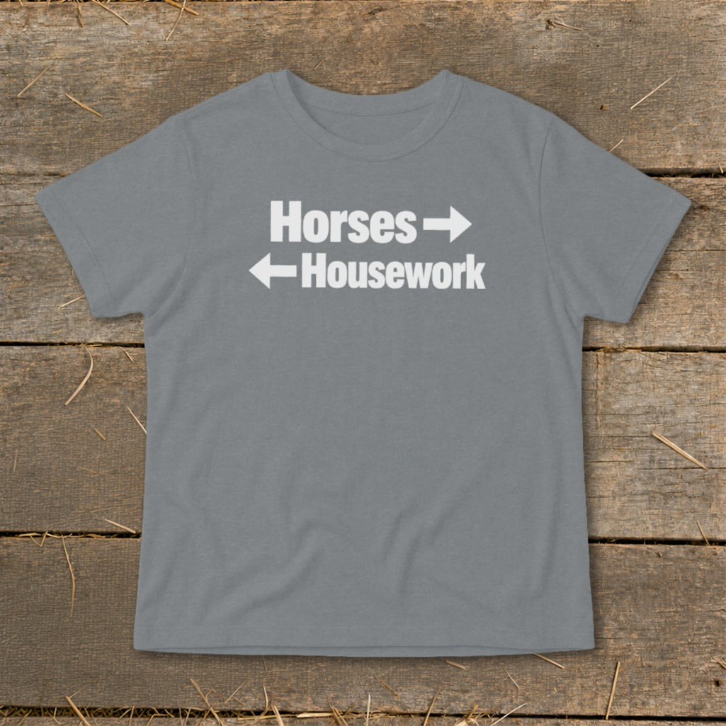 A grey short-sleeved t-shirt with white text reading Horses with a right arrow above Housework with a left arrow, laid flat on a rustic wooden background.