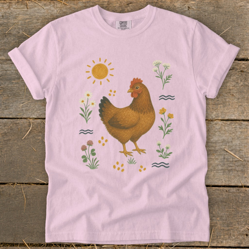 Pink t-shirt with a brown hen illustration surrounded by flowers, sun, and wavy lines.