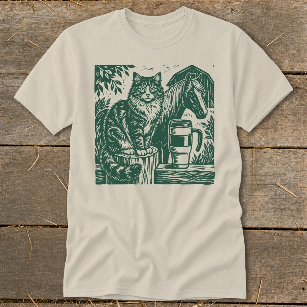Linocut design of a cat, horse, mug, and barn on a cream t-shirt.