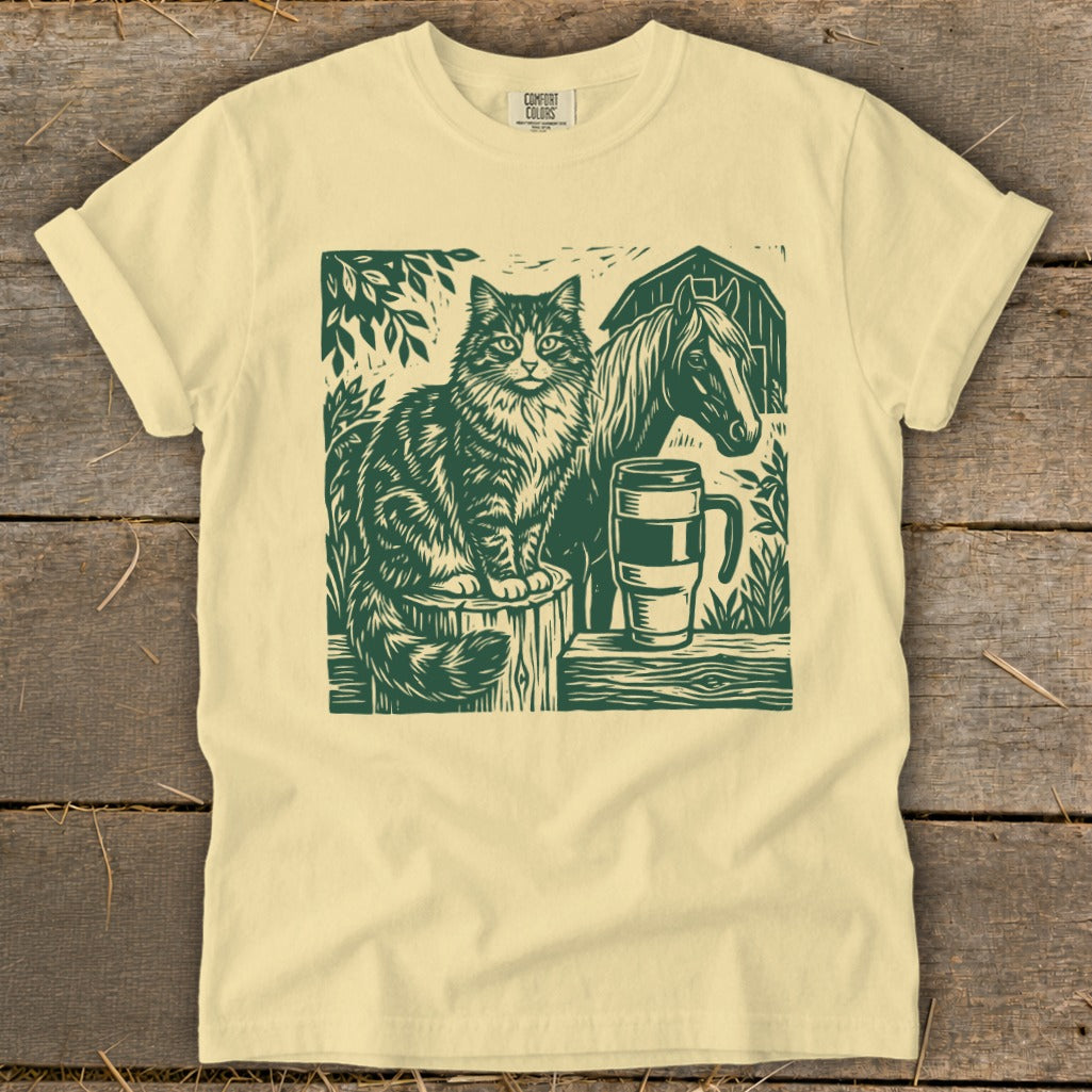 Green linocut print of a cat, horse, barn, and mug on a cream t-shirt.