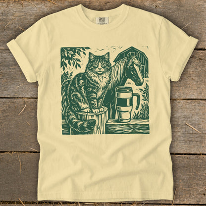 Green linocut print of a cat, horse, barn, and mug on a cream t-shirt.