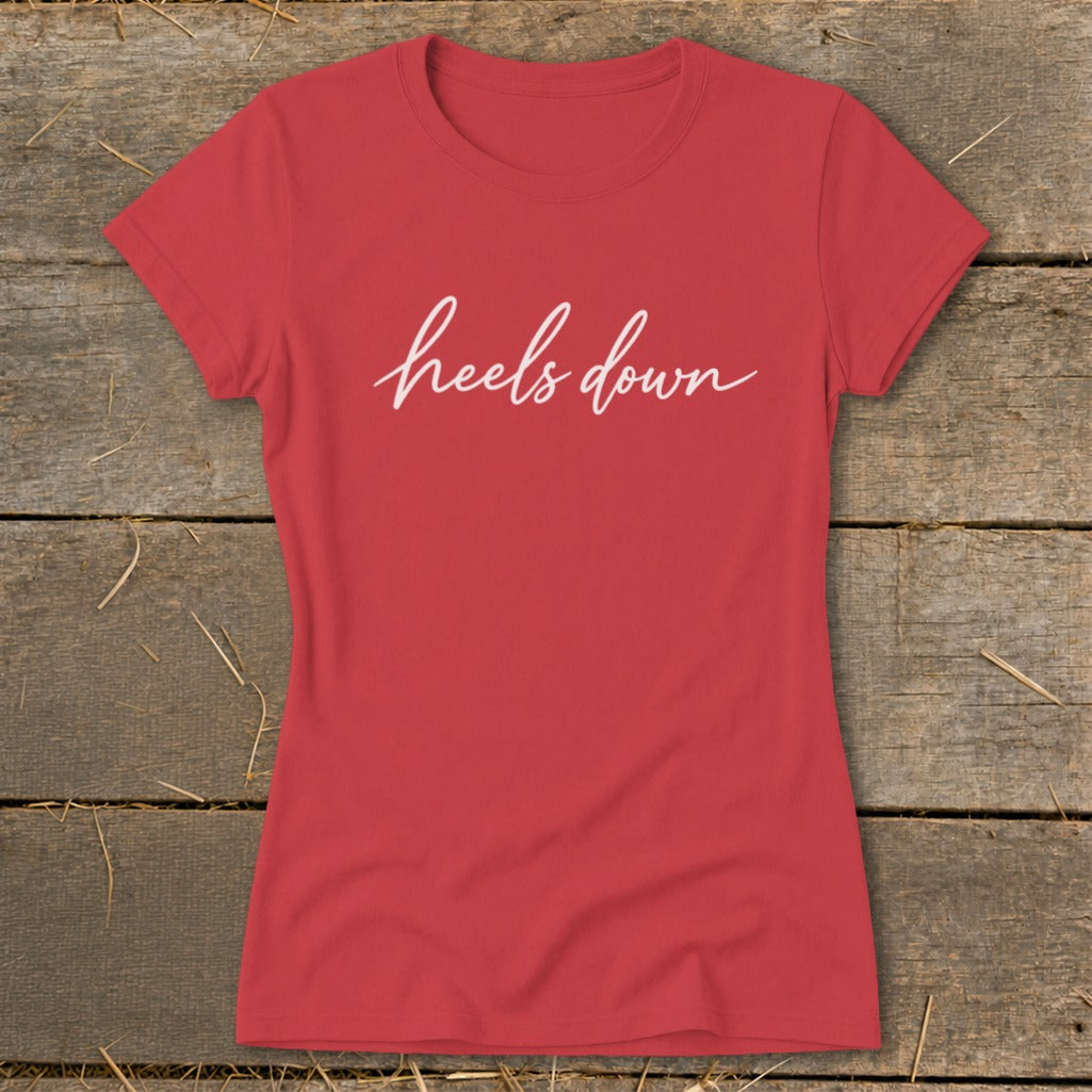 Red t-shirt with white cursive text reading "heels down" on rustic wood background.