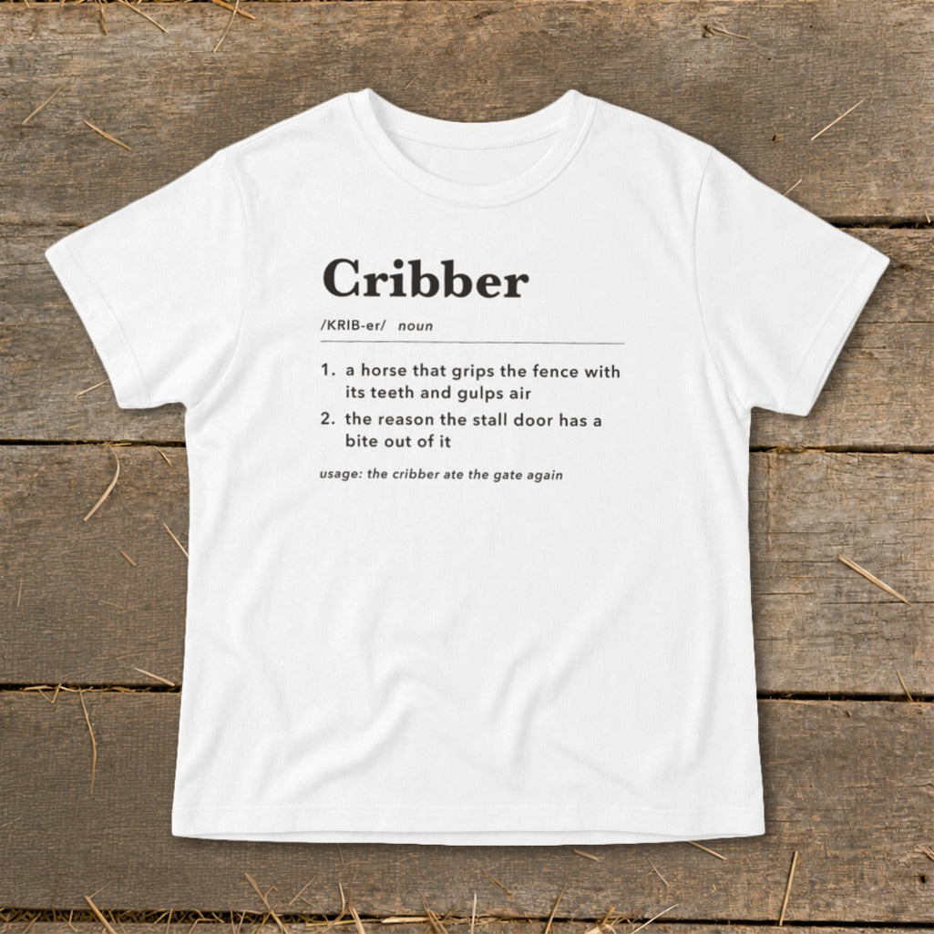 A white t-shirt featuring dictionary-style text defining the word Cribber on a wooden surface.