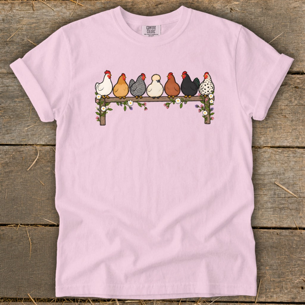 Light pink t-shirt featuring an illustration of seven colorful chickens sitting on a flower-adorned wooden fence.