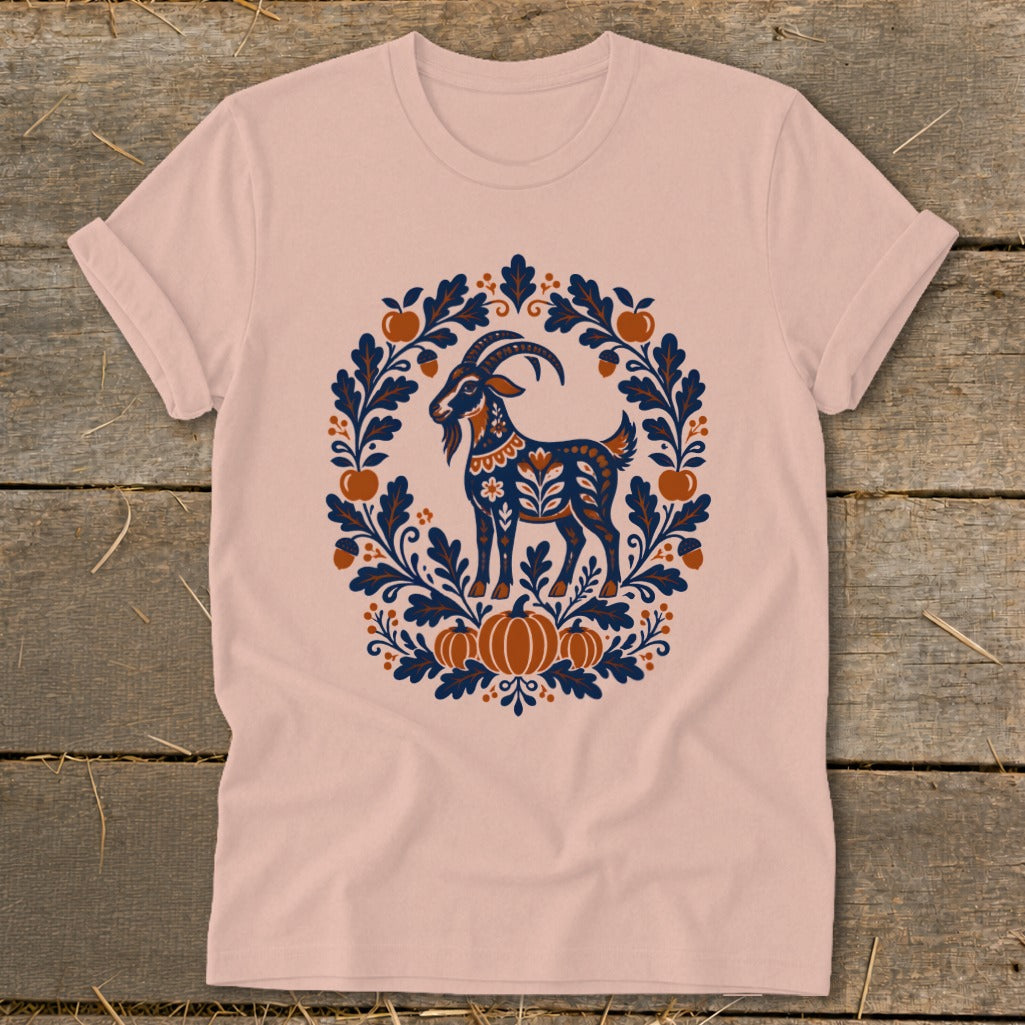 A peach-colored t-shirt with a folkloric illustration of a goat framed by autumn leaves and pumpkins.