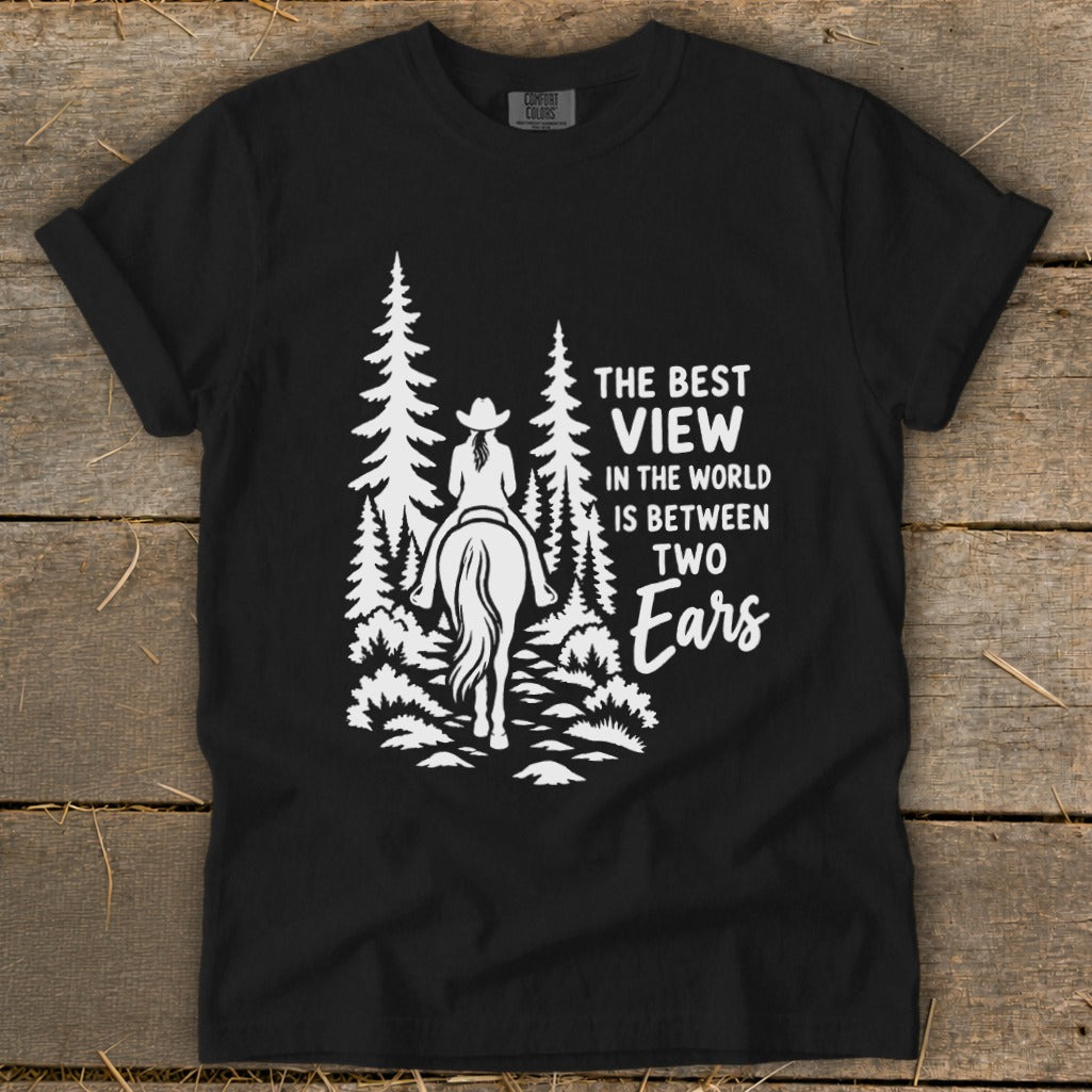 A black t-shirt featuring a white graphic of a horseback rider in the woods and text reading, "The best view in the world is between two ears."