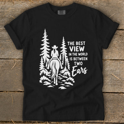 A black t-shirt featuring a white graphic of a horseback rider in the woods and text reading, "The best view in the world is between two ears."