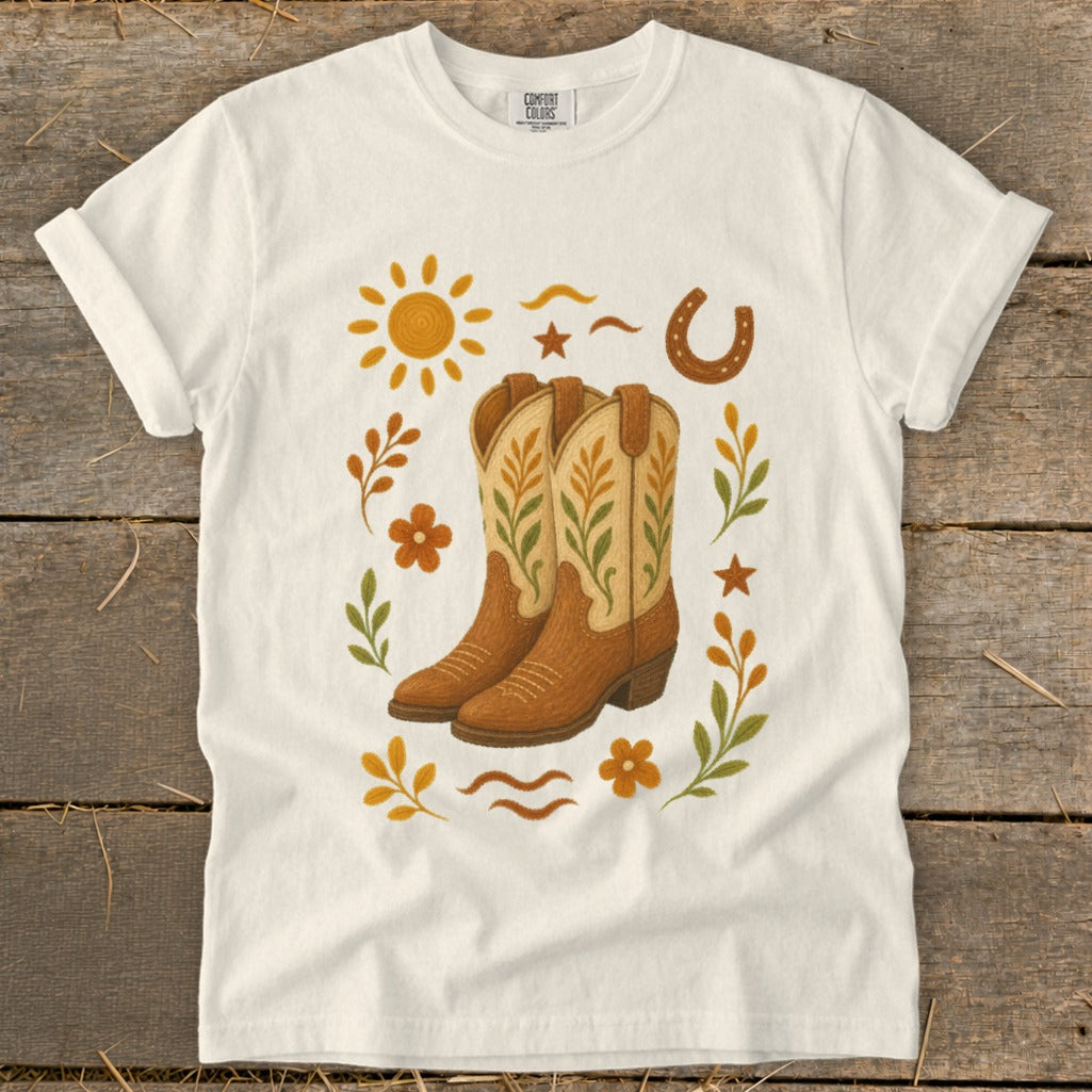 White t-shirt with a graphic of cowboy boots, sun, horseshoe, and foliage.