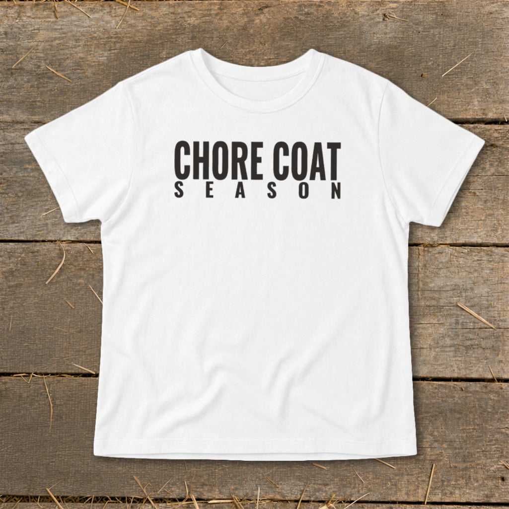 White t-shirt with black text "CHORE COAT SEASON" on a wooden background.